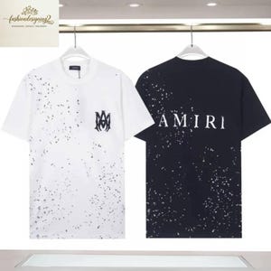 Könnte beinhalten: Zwei T-Shirts hängen an einem Kleiderbügel. Eines ist weiß mit schwarzem Spritzer und einem Logo, das andere ist schwarz mit weißem Spritzer und dem Wort "AMIRI". Beide haben kurze Ärmel.