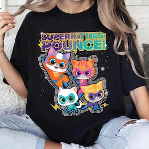 May include: A black t-shirt featuring a graphic of four cartoon cats dressed as superheroes. The text "SUPERKITTIES: POUNCE!" appears in gold and teal above the cats. The cats are orange, white, and yellow, wearing masks and capes.