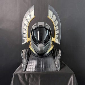Puede incluir: Un casco futurista con visera oscura y detalles plateados y dorados. El casco tiene grandes estructuras en forma de alas a cada lado. El casco se exhibe sobre una superficie negra.