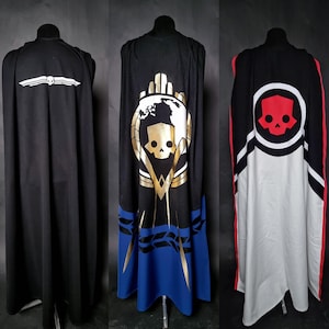 May include: Three black capes with unique designs. One features a white skull and wing emblem, another has a gold and blue emblem with a skull, and the third displays a red skull emblem with white and red accents.