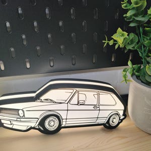 Könnte beinhalten: Schwarz-weiße Dekoration in Form eines Autos. Das Auto ist ein klassisches Schrägheck-Design mit detaillierten Umrissen der Fenster, Räder und Karosserie. Die Dekoration befindet sich auf einer Holzoberfläche, mit einer Topfpflanze im Hintergrund.