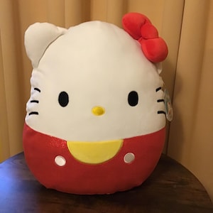 May include: A soft, round Hello Kitty cushion. The top half is white with black eyes, whiskers, and a yellow nose. A red bow sits on the left ear. The bottom half is red with a yellow accent and white dots.