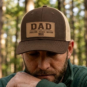 May include: Brown and tan trucker hat with a leather patch that reads "DAD" above the names "JACKSON - OLIVIA - MASON". The hat has a mesh back and a curved brim. The hat is worn by a person.