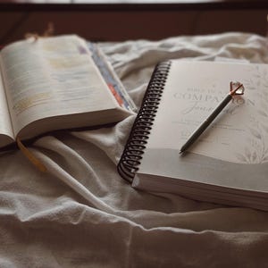 May include: An open book with colorful page markers, a spiral-bound journal titled BIBLE STUDY COMPANION JOURNAL with a pen resting on top, and a glass mug containing a floral-patterned liquid, all arranged on a white fabric surface.