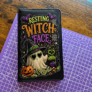 May include: A black rectangular notebook cover featuring a graphic print with the text "RESTING WITCH FACE" in orange and purple. The design includes a ghost wearing a witch hat and heart-shaped sunglasses, a jack-o'-lantern, money, and a cauldron.