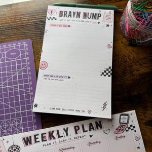 Op de afbeelding: Een wit papieren notitieblok met de titel BRAIN DUMP, zwarte tekst, roze accenten en een ruitpatroon. Het blok bevat secties voor notities en een checklist. Daaronder ligt een weekplanner met de tekst WEEKLY PLAN en dagelijkse kolommen.