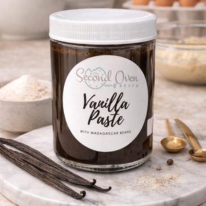 May include: A jar of vanilla paste with Madagascar beans. The jar is clear with a white lid and a label that reads "Second Oven Bakery Vanilla Paste." Vanilla beans and small gold spoons are on a marble surface.