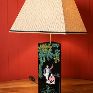 Peut inclure: Lampe noire rectangulaire avec abat-jour beige tissé. La base de la lampe présente une scène peinte de personnages sous un arbre. L'abat-jour est carré et bordé de noir. La lampe est sur une surface en bois contre un mur rouge.