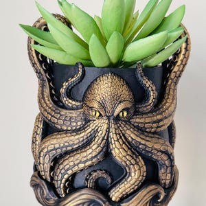 May include: A black and gold octopus-themed planter with a green succulent plant. The planter features an octopus design with detailed tentacles and a textured surface. The octopus has yellow eyes and is set against a black background.