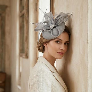 May include: A light gray fascinator hat featuring a structured circular base, intricate looped fabric ribbons, and delicate upright feathers. The headpiece is accented with two small, round, pearl-like beads at the center of the decorative arrangement.
