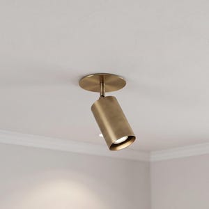 Könnte beinhalten: Ein zylindrischer Deckenstrahler aus Messing mit gebürsteter Metalloberfläche. Die Leuchte ist an einer runden Basisplatte montiert und verfügt über ein verstellbares Drehgelenk, mit dem der Leuchtenkopf nach unten ausgerichtet werden kann.