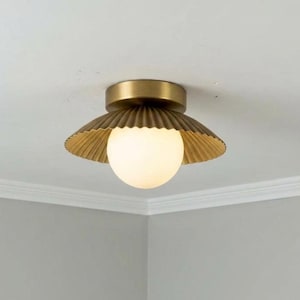 May include: A flush mount ceiling light fixture featuring a circular brass canopy and a wide, pleated, metallic brass shade. A spherical, frosted white glass globe sits centrally beneath the shade, emitting a soft, warm, diffused light.