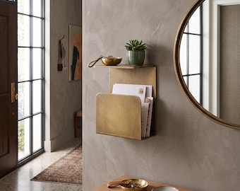 May include: A wall-mounted brass magazine rack with a flat top shelf holding a small green succulent in a ceramic pot and a small brass bowl. The rack contains several white papers and envelopes. The finish is a brushed metallic gold tone.