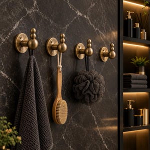 Puede incluir: Cuatro ganchos de pared de latón cepillado con diseño redondeado y escalonado, montados en una pared de mármol oscuro. Los ganchos sostienen una toalla gris oscuro de tejido de gofre, un cepillo de baño de madera con cuerda y una esponja.
