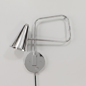 Pode incluir: Um candeeiro de parede em cromo polido com um abajur cónico fixado a um braço de estrutura retangular. A luminária inclui uma placa de montagem circular na parede com um pequeno interruptor e um cabo de alimentação preto na base.