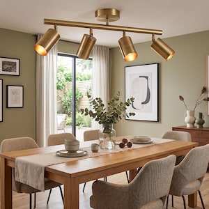 Puede incluir: Una lámpara de techo de cuatro luces con acabado en latón cepillado, que presenta focos cilíndricos montados en una barra horizontal. Las luces están orientadas hacia abajo, emitiendo un brillo cálido desde el interior de cada pantalla.