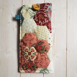 May include: A rectangular fabric case featuring a patchwork of hexagonal fabric pieces in shades of burgundy, cream, and sage green with floral and geometric patterns. The case includes a single wooden button closure at the top edge.