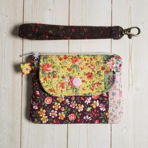 May include: A fabric pouch featuring a yellow floral flap with a pink snap button, a dark purple floral main body, and a light pink floral side panel. A matching dark brown floral wrist strap with a metal lobster clasp is included above the pouch.