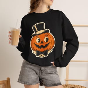 May include: A black long-sleeve sweatshirt featuring a graphic of an orange jack-o'-lantern wearing a black top hat and a black bow tie. The pumpkin has a wide, smiling face with large, expressive eyes in a classic cartoon style.