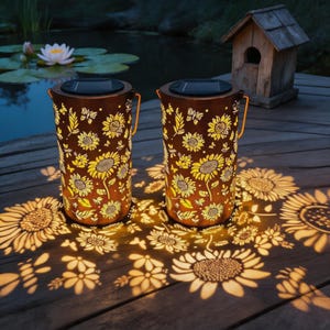 May include: Two cylindrical metal solar lanterns with a bronze finish, featuring intricate cut-out patterns of sunflowers and butterflies. The lanterns emit a warm, glowing light that projects large, detailed sunflower silhouettes onto the surface.