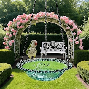 Könnte beinhalten: Ein Vogelbad aus grünem Glas mit gewelltem Rand und geprägten Blumenmustern, das an drei schwarzen Metallketten hängt. Ein kleiner grüner und grauer Papagei mit rotem Schnabel steht auf dem Rand der Schale, die mit klarem Wasser gefüllt ist.