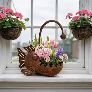 Könnte beinhalten: Ein rostfarbener Metall-Pflanzkübel in Form eines Anglerfisches, gefüllt mit rosa Tulpen, violetten Hyazinthen und kleinen weißen Blumen. Zwei braune geflochtene Hängekörbe mit rosa Geranien hängen an beiden Seiten des Fensterrahmens.