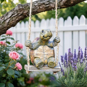 May include: A decorative turtle figurine with a textured brown and green shell, sitting on a small wooden swing suspended by two thick jute ropes. The turtle has a smooth, light-colored plastron and is positioned in a garden setting with flowers.