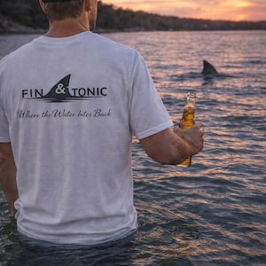 May include: A white t-shirt with the text "FIN & TONIC" and a shark fin graphic. The shirt also has the phrase "Where the Water bites Back". A person stands in the water holding a beer bottle, with a shark fin visible in the distance.