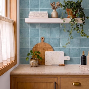 May include: A kitchen wall featuring square, glossy, teal-colored tiles. A white floating shelf holds three books titled The Farmhouse Kitchen, Slow Living, and Nature's Larder, a beige vase with dried flowers, and a potted ivy plant.