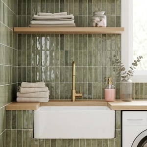 Könnte beinhalten: Ein modernes Badezimmer mit einem weißen Bauernhauswaschbecken, einer goldenen Armatur und grün gefliesten Wänden. Holzregale beherbergen gefaltete Handtücher und ein Glas. Ein rosa Seifenspender und eine Vase mit Grün.