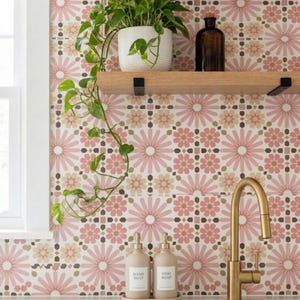 Op de afbeelding: Een keuken met een gouden kraan, twee beige zeepdispensers en een dienblad. Een witte pot met een groene plant staat op een houten plank, met een bruine fles. De achterwand heeft een bloemenpatroon in roze, bruin en wit.