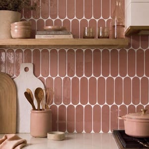 May include: A kitchen backsplash featuring vertical, pill-shaped ceramic tiles in a dusty rose colour with white grout. The tiles are arranged in a repeating pattern, creating a smooth, glossy surface that covers the wall behind the wooden shelf.