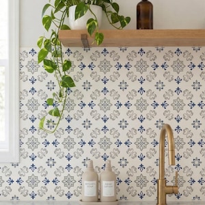 May include: A kitchen scene featuring a gold faucet, a patterned backsplash with blue and gray floral designs, and two beige pump bottles labeled "Kitchen Soap" and "Hand Lotion." A trailing green plant and a brown bottle sit on a wooden shelf.