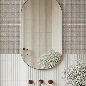 Peut inclure: Un miroir ovale à cadre sombre est suspendu au-dessus d'un lavabo blanc. Le mur est carrelé de petits carreaux carrés et de longues et fines tuiles dans des tons de gris et de blanc. Robinetterie en bronze et vase de fleurs blanches.