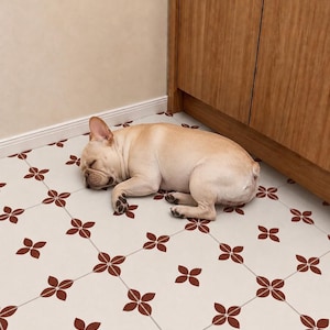 May include: A fawn-colored French Bulldog sleeping on a white floor with a repeating pattern of small, dark red four-petaled flower shapes. The dog is curled up in a corner next to a light brown wooden cabinet with vertical wood grain.
