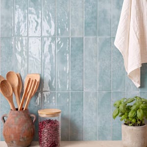May include: A kitchen scene featuring light blue, glossy, rectangular wall tiles. A terracotta pot holds wooden spoons, a glass jar contains red beans, and a small concrete planter holds a green basil plant. A white cloth hangs on the wall.