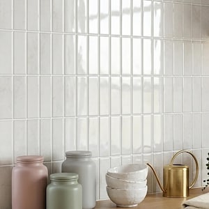 Pode incluir: Uma parede é coberta por azulejos de cerâmica brancos retangulares dispostos verticalmente. Abaixo, uma superfície de madeira abriga itens de cozinha: um pote rosa, um pote verde, um pote cinza, uma pilha de tigelas brancas e um regador dourado.