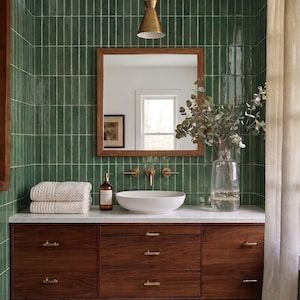 May include: A bathroom vanity featuring a dark wood cabinet with brass hardware, a white vessel sink, and a marble countertop. The wall is covered in vertical green tiles, and a square wooden mirror hangs above the sink with a brass pendant light.