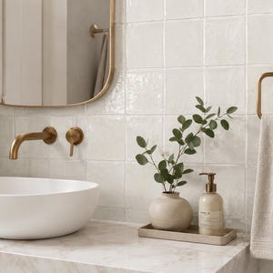 Puede incluir: Un &aacute;rea de lavabo de ba&ntilde;o con un lavabo blanco sobre encimera, un grifo de pared de lat&oacute;n y un espejo con marco de lat&oacute;n. Un jarr&oacute;n de cer&aacute;mica beige con ramas de eucalipto y un frasco con dosificador descansan sobre una bandeja rectangular.