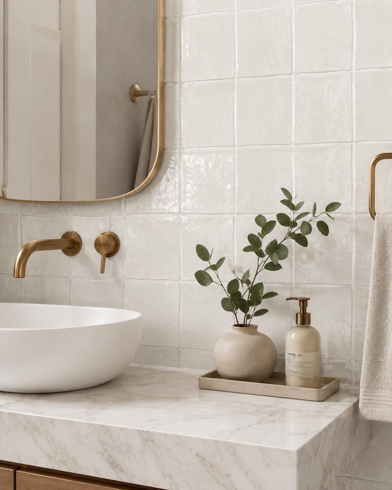 Puede incluir: Un &aacute;rea de lavabo de ba&ntilde;o con un lavabo blanco sobre encimera, un grifo de pared de lat&oacute;n y un espejo con marco de lat&oacute;n. Un jarr&oacute;n de cer&aacute;mica beige con ramas de eucalipto y un frasco con dosificador descansan sobre una bandeja rectangular.