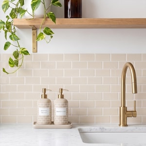 May include: A beige ceramic hand soap and dispenser set with gold-colored pumps, sitting on a matching tray. The labels read "HAND SOAP" and "DISPENSER". The set is on a white countertop with a gold faucet and beige brick-style tile backsplash.