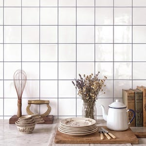 May include: A kitchen scene featuring a stack of floral patterned plates, a white enamel teapot with a blue rim, a wire whisk, a brass tool, a glass jar with dried flowers, and three vintage books arranged on a marble countertop with white tiles.
