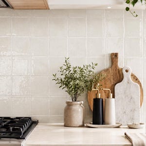 May include: A kitchen counter featuring a beige ceramic vase with a green plant, two cylindrical soap dispensers in black and beige with gold pumps on a tray, a round wooden cutting board, and a white marble cutting board against tiled walls.
