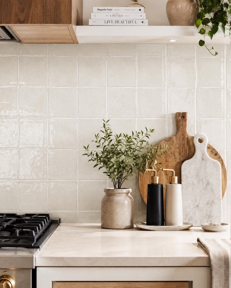 Puede incluir: Una encimera de cocina con un jarr&oacute;n de cer&aacute;mica beige con una planta verde, dos dispensadores de jab&oacute;n cil&iacute;ndricos en negro y beige con bombas doradas sobre una bandeja, una tabla de cortar de madera y una tabla de m&aacute;rmol blanco.