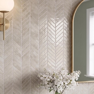 May include: A bathroom wall featuring a light beige herringbone tile design. A gold-framed mirror and a brass wall light with a white globe are present. A vase of white flowers is in front of the mirror.