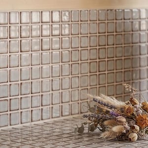 May include: A wall and surface covered in small, square, glossy light gray tiles with dark grout lines. A small, dried floral arrangement featuring lavender, wheat, and various seed pods rests on the tiled surface in the lower right corner.