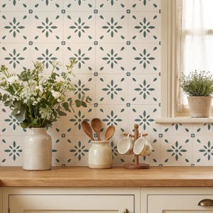 May include: A kitchen interior with patterned tiles, a wooden worktop, and cream cabinets. A vase of white flowers, wooden spoons, and mugs on a wooden stand are visible. A small potted plant sits on the windowsill, adding a touch of greenery.