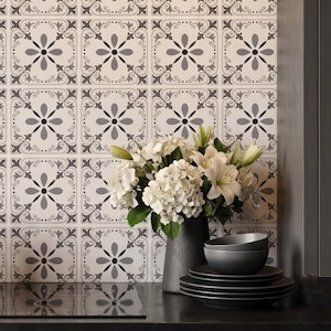 May include: A still life featuring a bouquet of white flowers in a dark gray vase, with matching plates and bowls. The background is a patterned tile wall in white, gray, and black. The scene is set on a dark countertop.