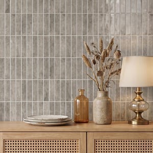 Può includere: Scena d'interni decorativa con una parete piastrellata grigia con piastrelle rettangolari verticali. Un mobile in legno con un design intrecciato contiene una lampada, vasi e piatti. La lampada ha una base dorata e un paralume color crema. Piante secche in un vaso.