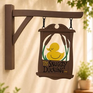 Op de afbeelding: Een bruin houten wandbord met een rechthoekig hangbord. Het bord is voorzien van een illustratie van een gele badeend, groene lisdoddeplanten en de tekst The Snuggly Duckling in zwarte letters op een bruine achtergrond.